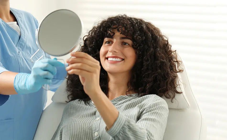 Patient smiling in dental chair viewing teeth in mirror after whitening treatment at cosmetic dentist