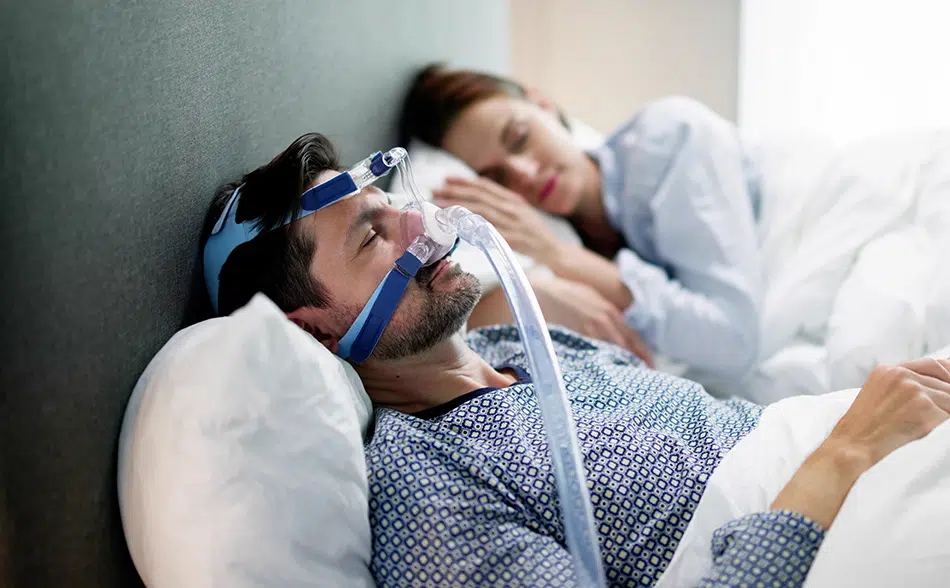 Man wearing CPAP mask sleeping beside partner, illustrating sleep apnea treatment for better oral health